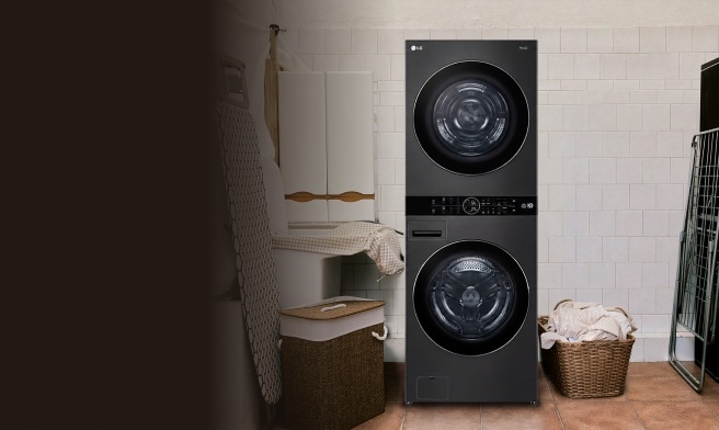 A black LG WashTower® with the dryer on top and the washer below, set up in a white-tiled laundry room next to wicker laundry baskets, an ironing board and a drying rack.