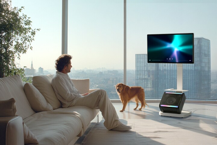 Man relaxing with a dog in a high-rise living room while watching content on LG Smart Monitor Swing mounted on Swing Stand and Swing Stand Plate, with XBOOM Stage 301 on the base and a remote control placed on the plate