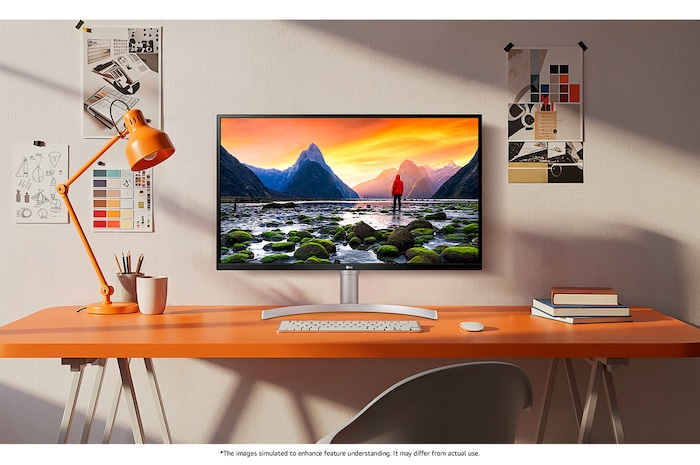 The LG Monitor 32UN650K in a white/silver finish, featuring UHD 4K resolution, displays a brilliant landscape image in a stylish, creative setting on an orange desk.