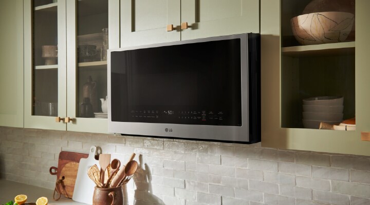A silver countertop microwave sits in a modern kitchen with a gray stone backsplash, a sink, and a built-in oven.