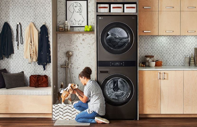 Black steel LG WashTower in a mudroom laundry space with light wood cabinets, gray tile backsplash, bench, and wall hooks.