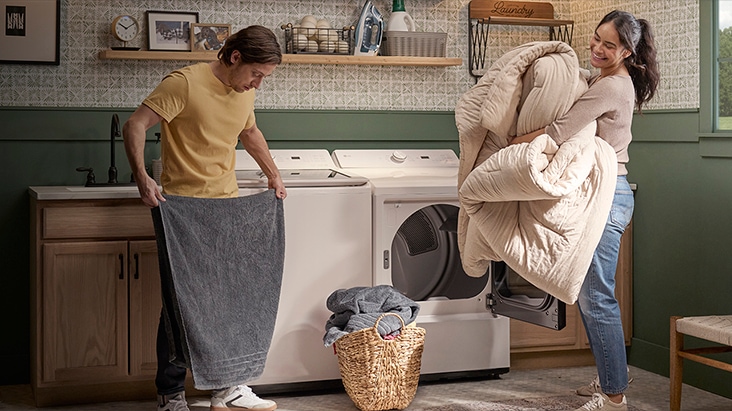 Person holding a gray towel beside an LG washer and dryer while another person loads a comforter into the dryer