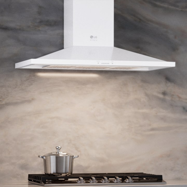 White LG Studio wall-mount range hood with built-in light above a gas cooktop and marble backsplash.