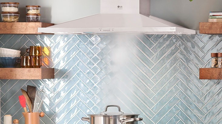 LG Studio stainless steel wall mount range hood extracting steam from a pot over a blue herringbone tile backsplash.