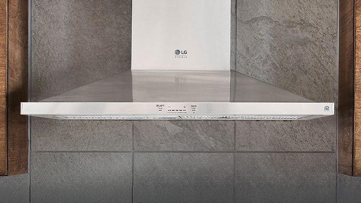 LG Studio stainless steel wall-mount range hood with touch controls mounted against a grey stone tile wall.