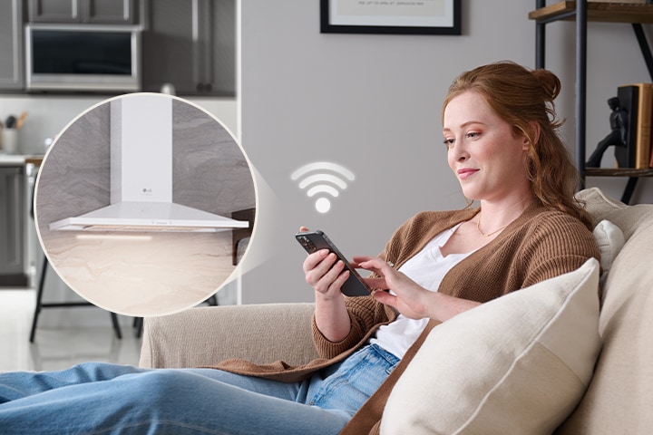 A woman uses a smartphone to remotely control a white LG wall-mount range hood via Wi-Fi.