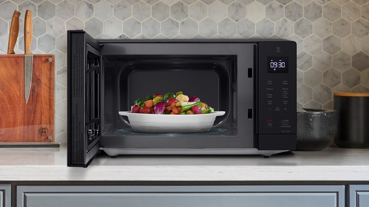 Open black countertop microwave with a white dish of roasted vegetables inside on a kitchen counter.