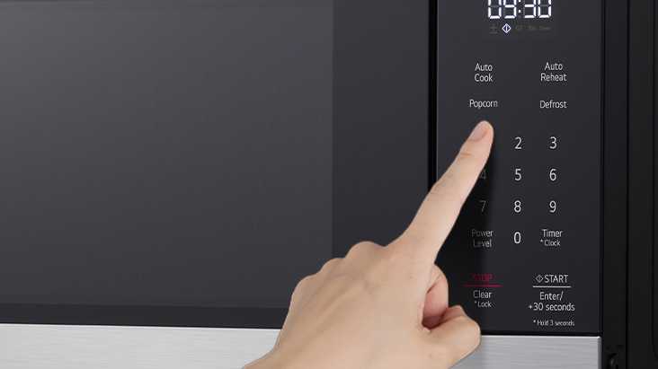 A hand pressing the popcorn button on a black microwave control panel with a digital display.