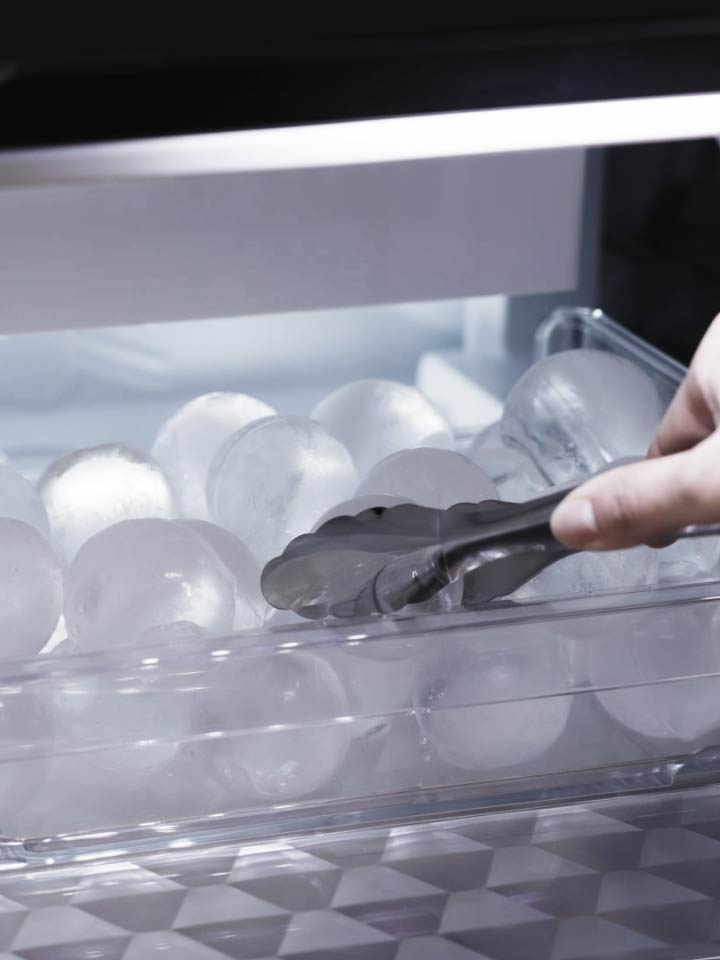 Tongs picking up a round ice ball from a transparent tray inside a freezer