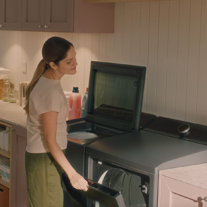 A woman opens the door of a modern dark gray front-load dryer next to a matching top-load washing machine.