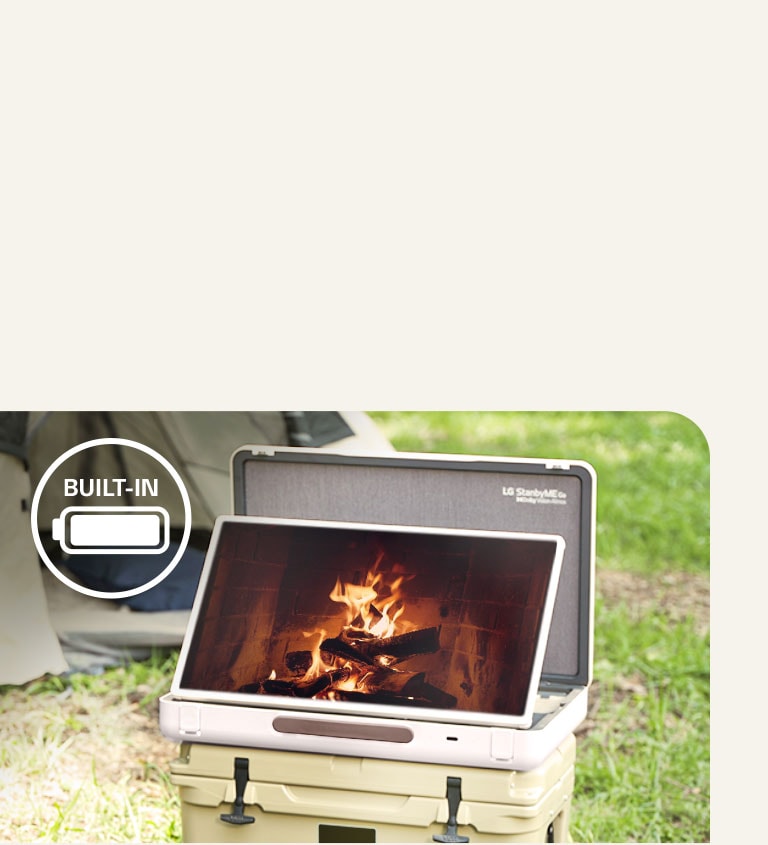 The LG StanbyME Go is placed in front of a tent, and the screen displays one of the relaxing themes--a fireplace. On the left top corner, built-in battery icon is shown.