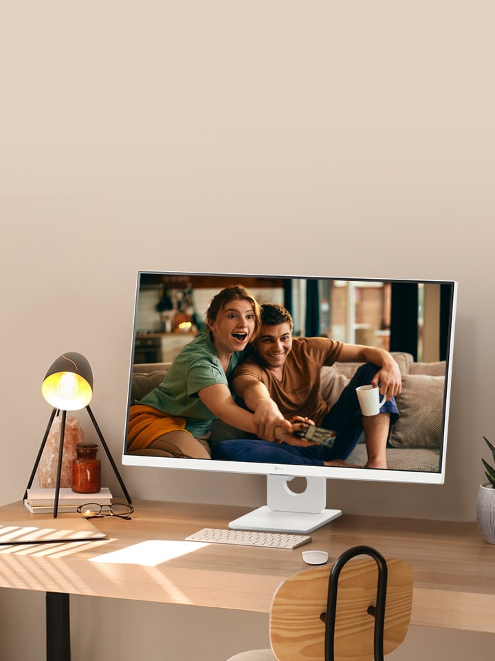On the desk by the sunlit window, there is a smart monitor displaying a movie on its screen. Beside the monitor, there is a lamp and a potted plant. On the desk, there is a keyboard and a mouse.
