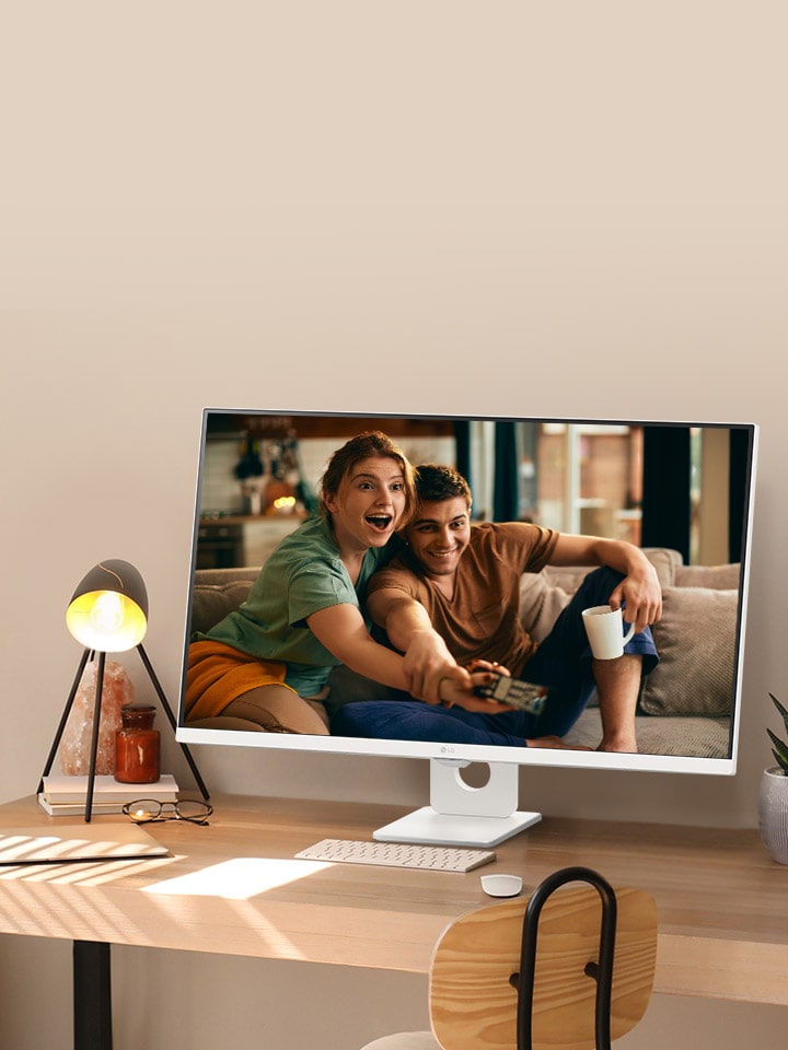 On the desk by the sunlit window, there is a smart monitor displaying a movie on its screen. Beside the monitor, there is a lamp and a potted plant. On the desk, there is a keyboard and a mouse.