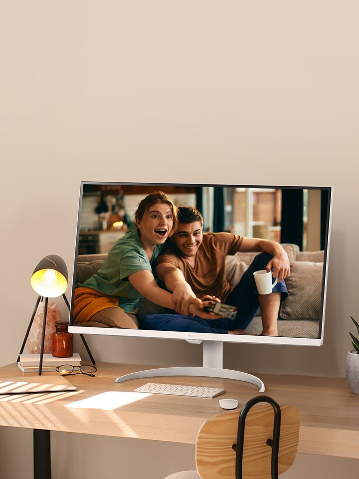 On the desk by the sunlit window, there is a smart monitor displaying a movie on its screen. Beside the monitor, there is a lamp and a potted plant. On the desk, there is a keyboard and a mouse.