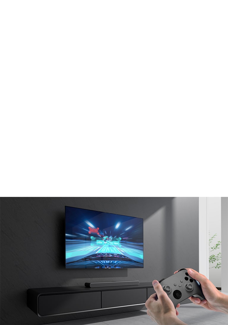 The sound bar is placed on the cabinet and racing game scene is shown on the TV coneected to the sound bar. A game console is on the bottom right side of the picture hold by two hands. 