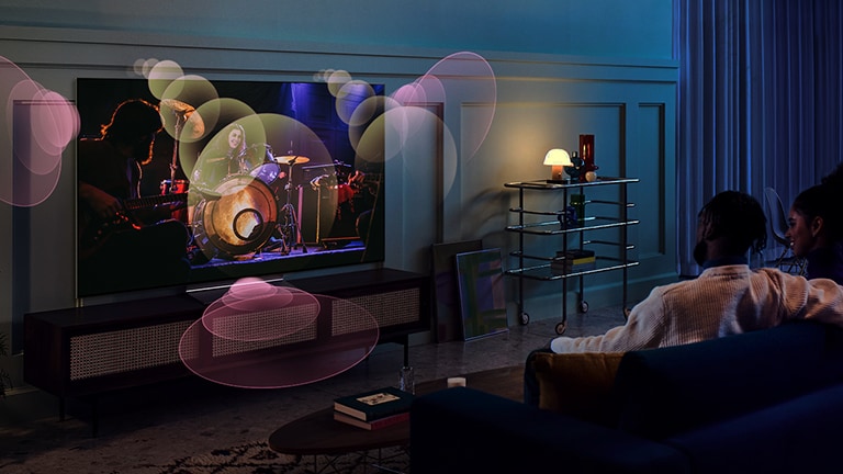 People sit on a couch watching a concert with bubbles depicting surround sound around them.