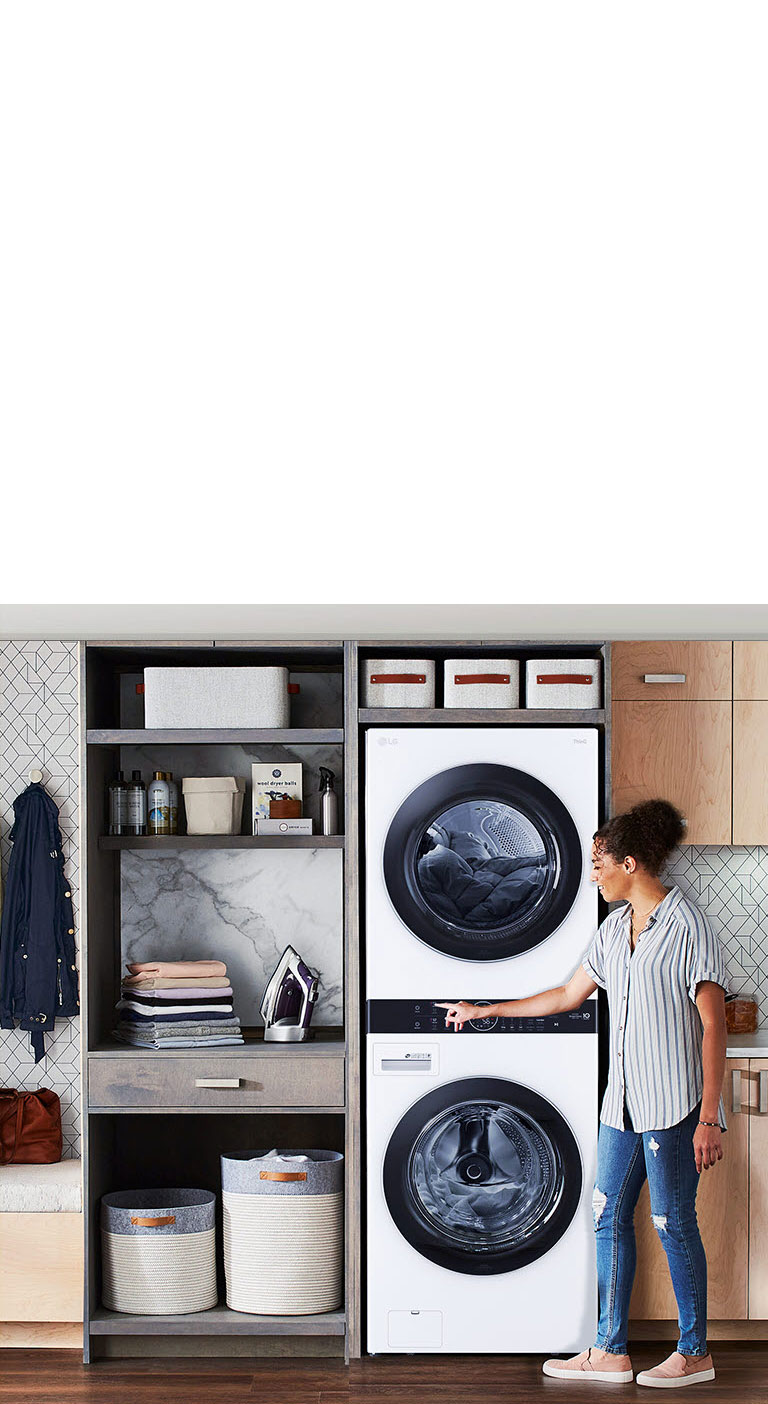 Modern laundry room with stacked washer-dryer, cabinets, and bench on left, woman interacting with control panel.