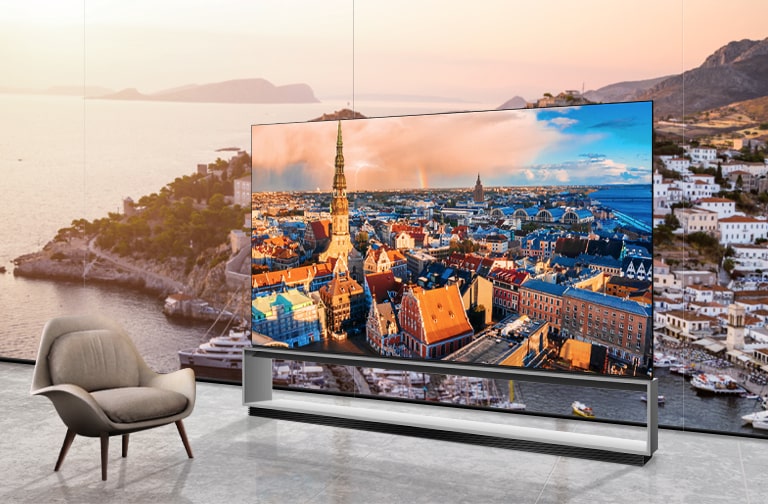 Room overlooking a seaside town. A TV is installed in front of the window showing a view of a city skyline. There is a chair next to the TV.