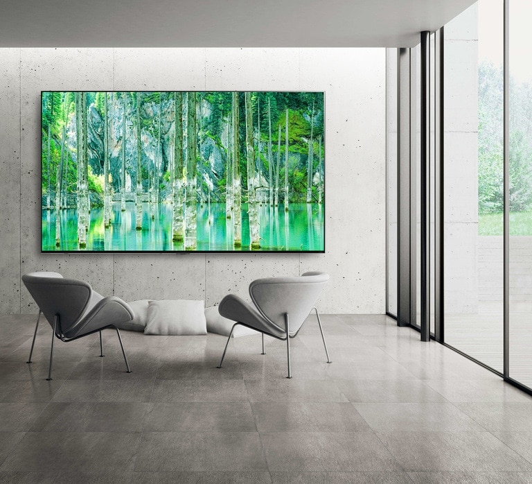 Image of a large-screen LG QNED MiniLED TV mounted against a grey, bare concrete wall. Two chairs are arranged in front of the TV and the screen shows a green natural scene.