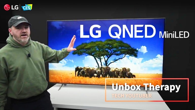 Tech YouTuber Unbox Therapy standing in front of an LG QNED MiniLED TV. The screen shows an image of a herd of elephants surrounding a single tree.