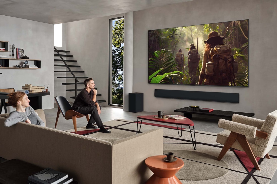 Vue large d’un salon à l’intérieur d’une maison haut de gamme, un homme regarde un film sur son LG OLED TV fixé au mur. La conception du téléviseur s’intègre élégamment au mur et à la décoration intérieure.