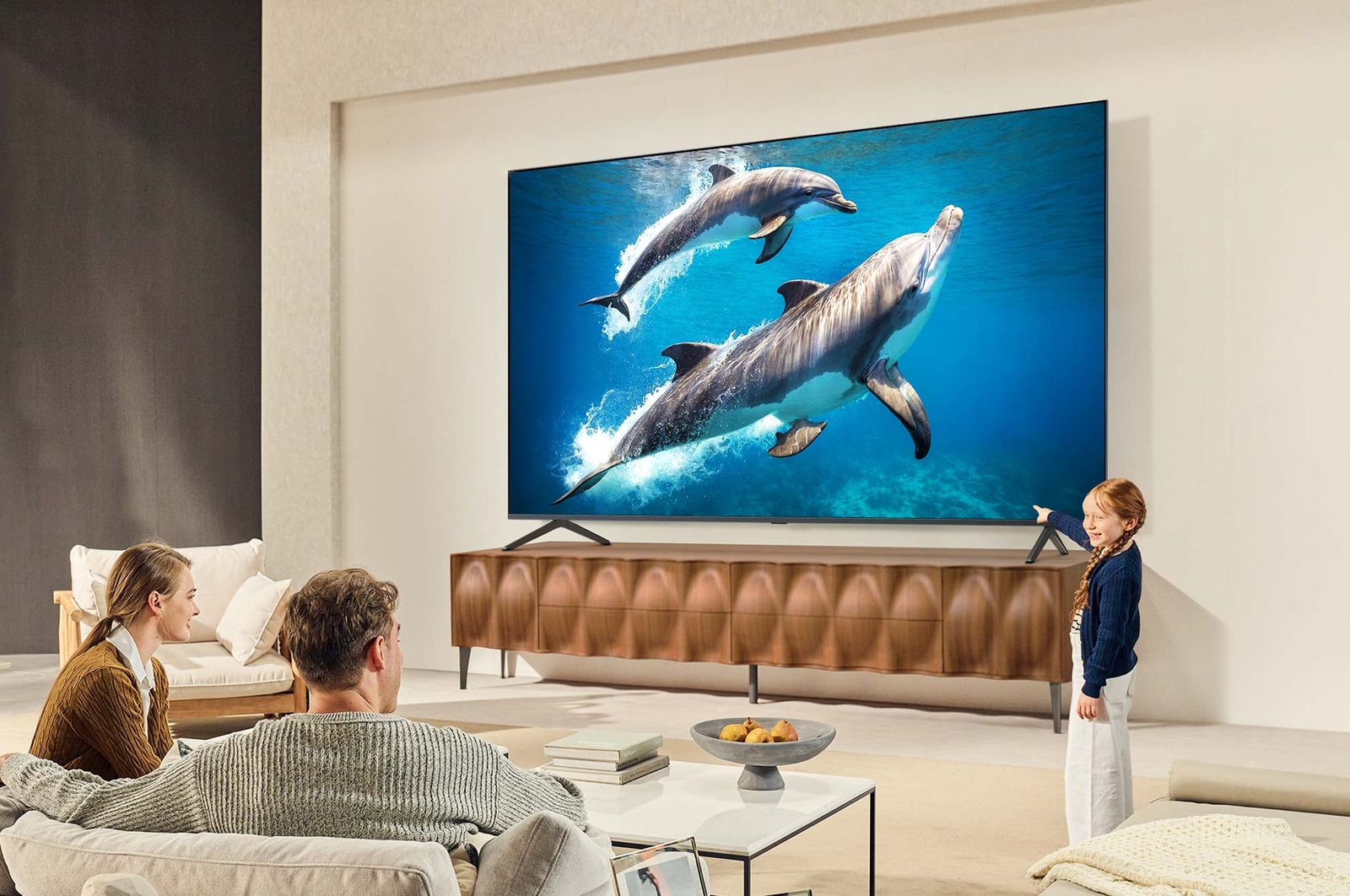 Une famille est assise sur un canapé face à un LG QNED TV placé au-dessus d’un meuble TV. Une petite fille pointe du doigt un écran représentant deux dauphins. Le titre explique comment profiter de la haute résolution sur un écran géant.