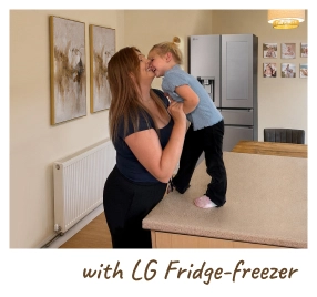 Une mère tient son bébé dans une cuisine moderne, debout à côté d’un réfrigérateur ouvert. [with LG Fridge-freezer]