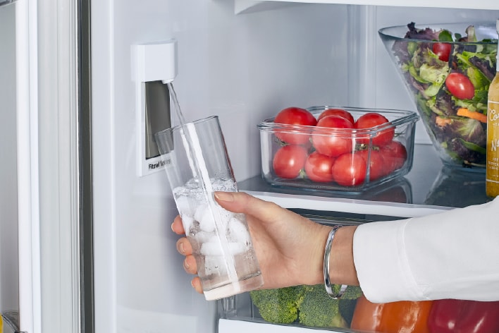 Une main remplit un verre de glaçons avec le distributeur d'eau interne d'un réfrigérateur blanc.