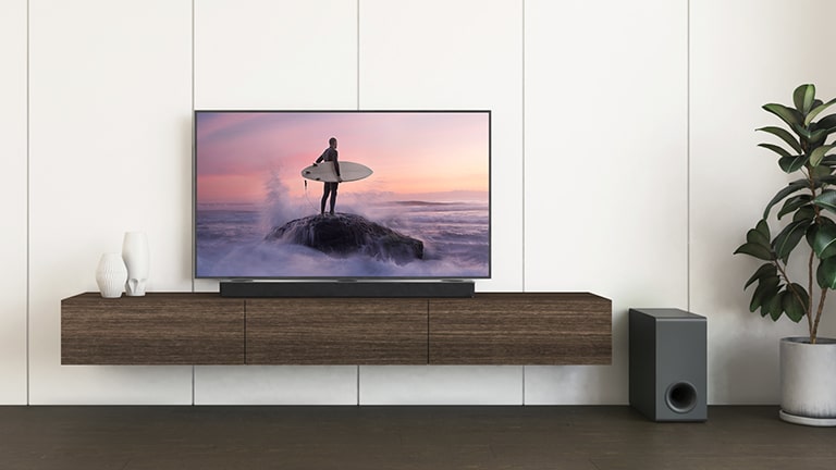 Un téléviseur de LG et une barre de son de LG sont placés sur une étagère brune, et un caisson de basses est au sol. Le téléviseur montre un surfer debout sur un rocher.