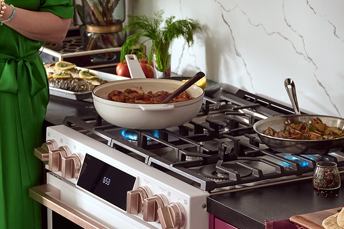 Cuisinière à gaz blanche avec boutons dorés, poêles sur le feu et plats cuisinés sur un plan de travail.