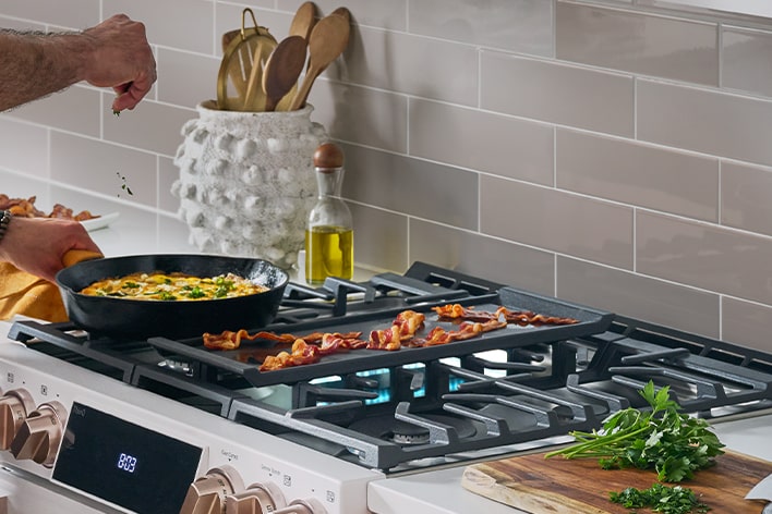 Cuisinière à gaz blanche avec bacon sur une plaque chauffante et omelette dans une poêle en fonte.