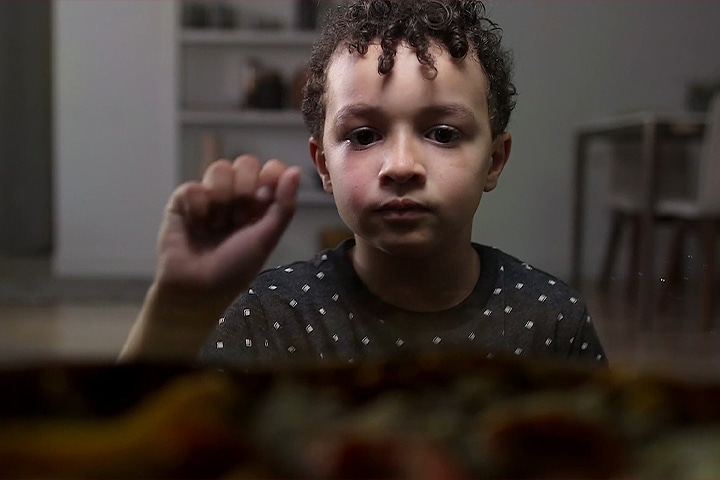 Jeune garçon aux cheveux bouclés regardant attentivement une pizza au premier plan.