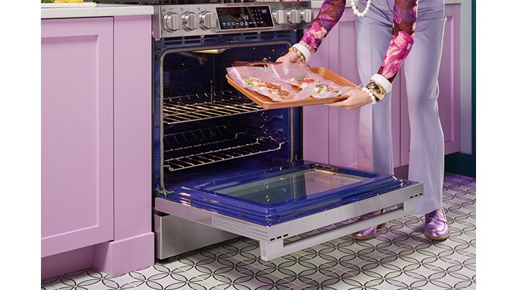 Une femme place une plaque de cuisson avec des plats dans un four encastré en acier inoxydable, entouré d'armoires roses.