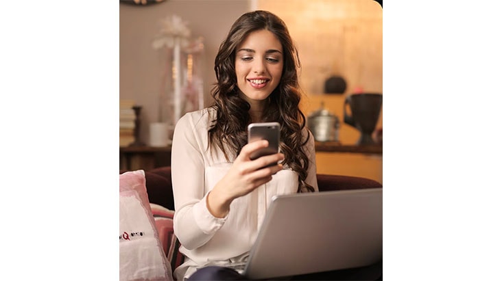 Femme souriante assise avec un ordinateur portable, utilisant son smartphone dans un salon chaleureux.