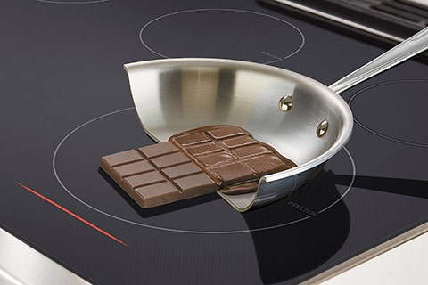 Une barre de chocolat et une poêle sont placées sur la surface de cuisson à induction, démontrant l’aspect sécuritaire de la surface froide au toucher même pendant la cuisson.