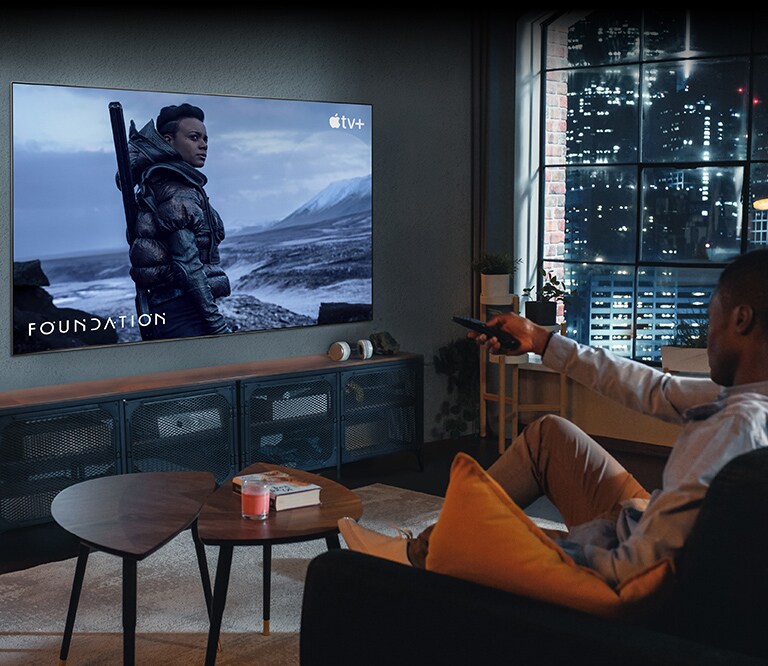 Un homme est assis sur le canapé et regarde la télévision. L'homme tient une télécommande à la main et la télévision affiche une scène de la série Fondation sur Apple TV+.