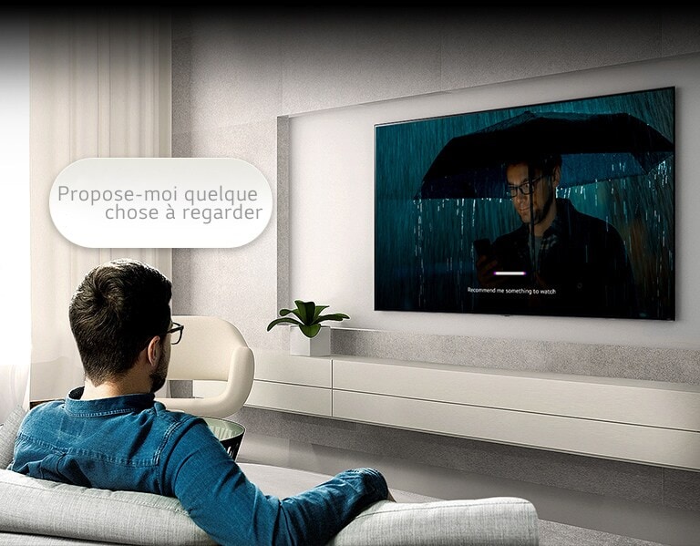 Un homme est assis sur un canapé et fait face à un téléviseur situé de l'autre côté. Une bulle de dialogue indiquant « Propose-moi quelque chose à regarder » flotte au-dessus de sa tête. À l'intérieur de l'écran, une vidéo d'un homme portant un parapluie est diffusée avec une interface utilisateur à reconnaissance vocale.