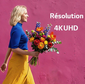 L’écran mobile StanbyME 2 Max LX6 de LG, doté de la résolution 4K UHD, montre une femme portant des vêtements colorés et tenant des fleurs qui passe devant un mur de rue violet, mettant en évidence la netteté et la précision de la résolution.