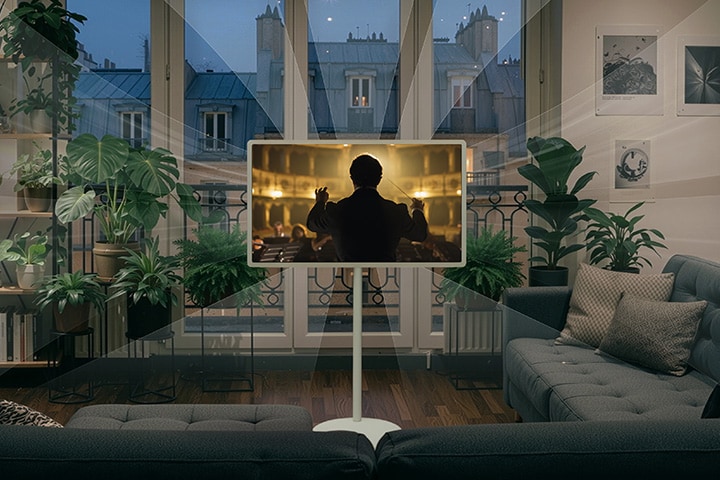 L’écran mobile StanbyME 2 Max LX6 de LG avec fonctionnalité Son IA Pro montre une scène d’orchestre dans une pièce confortable, sombre et remplie de plantes, avec des ondes sonores qui s’étendent vers une personne lisant sur un canapé, illustrant un son immersif qui remplit la pièce.
