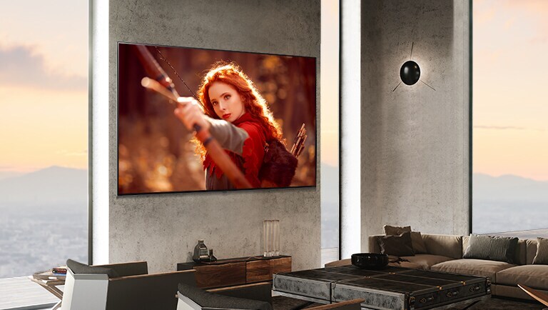 Un grand téléviseur fixé sur un mur de pierre grise à côté de très grandes fenêtres dans une pièce moderne. L’écran montre une femme rousse qui pointe une flèche d'arc vers la caméra.