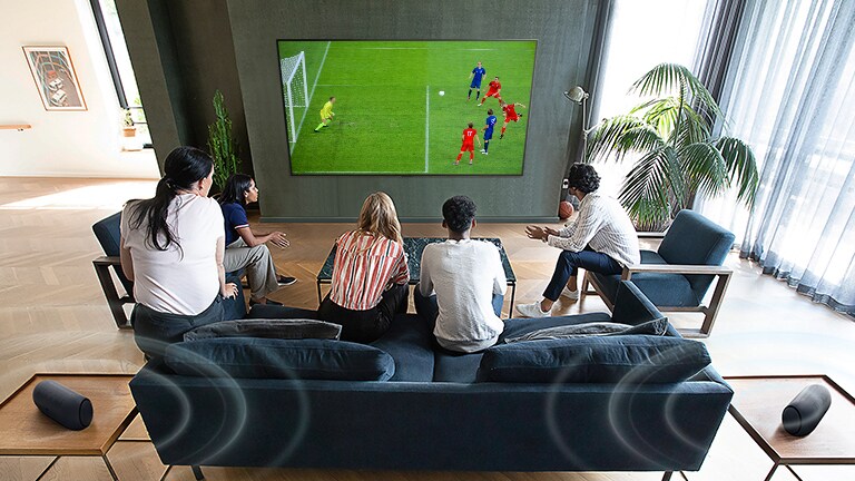 Cinq personnes rassemblées devant un téléviseur à écran plat fixé au mur et regardant une partie de soccer.