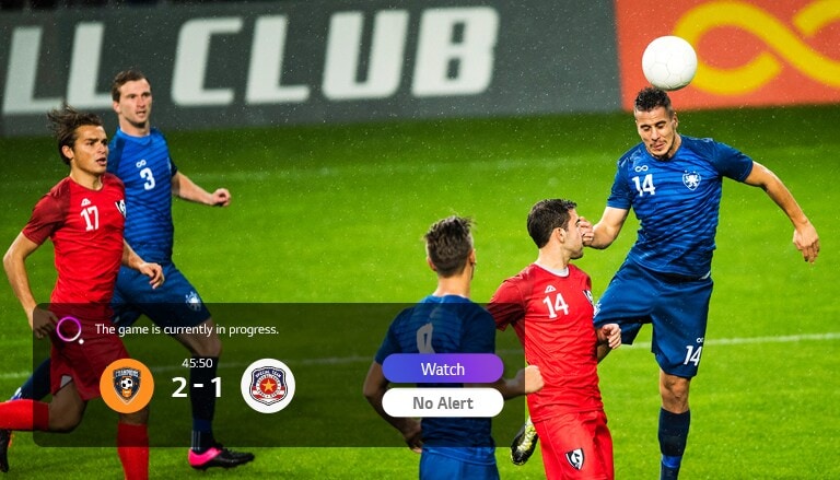 Un couple assis sur un canapé regarde un match de soccer sur un téléviseur qui affiche la notification d'un autre match de sport en bas à gauche.