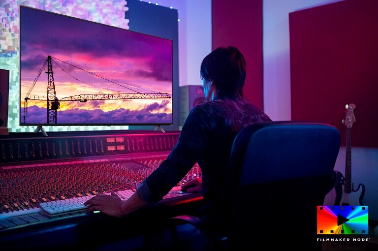 Un réalisateur de film est en train de modifier quelque chose en visionnant un grand téléviseur. L’écran de téléviseur affiche une grue à tour contre un ciel violet. Le logo du mode CINÉASTE est affiché en bas à droite.