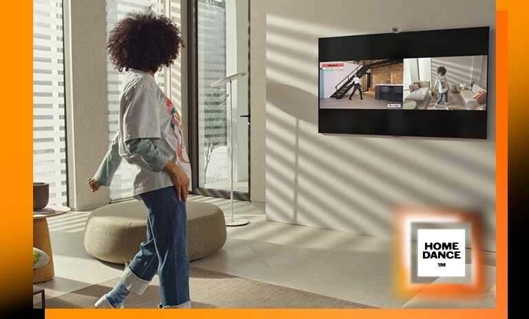 Une femme debout au milieu du salon danse et regarde la télé en même temps. Le téléviseur montre une scène de didacticiel de danse à gauche et reflète une femme qui danse à droite de l'image. Le logo de 1Million HomeDance est en bas à droite.