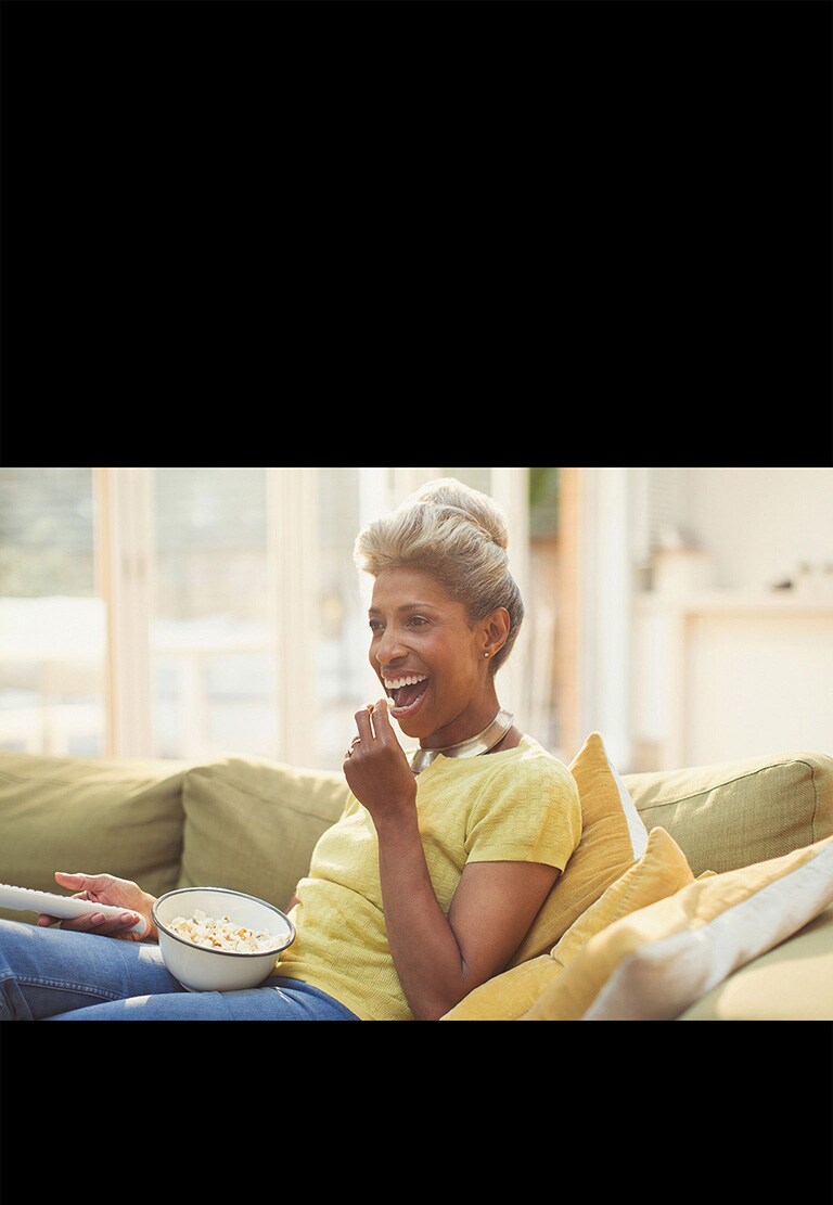 Une femme regarde la télévision, télécommande en main. Elle mange aussi du maïs soufflé.