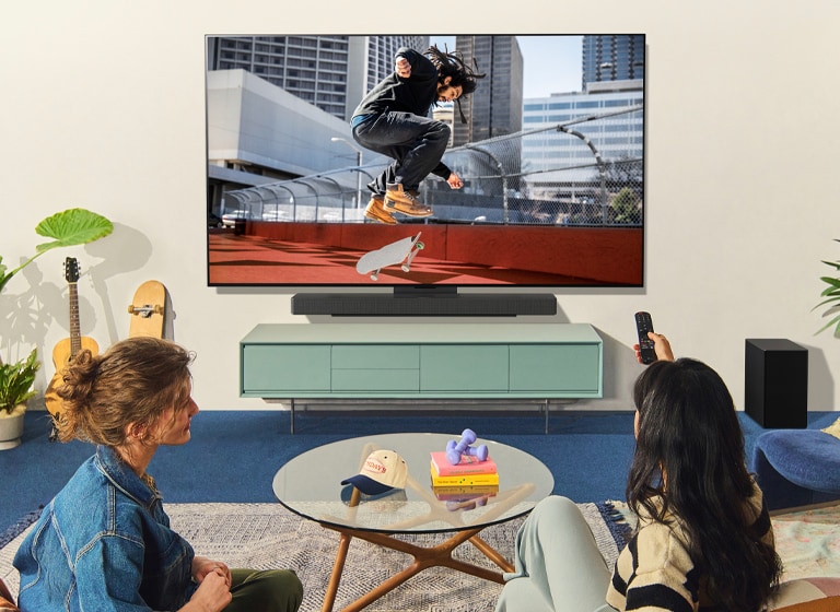 Zwei Frauen sitzen vor einem wandbefestigten LG TV auf einem Sofa in einem farbenfrohen Wohnzimmer und eine Frau zeigt mit der Fernbedingung auf einen TV-Bildschirm. 