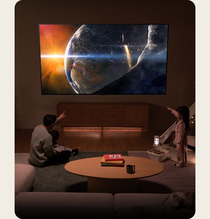 Eine Familie sitzt auf dem Boden eines schwach beleuchteten Wohnzimmers an einem kleinen Tisch und blickt zu einem LG-TV an der Wand, der die Erde aus dem Weltraum zeigt.