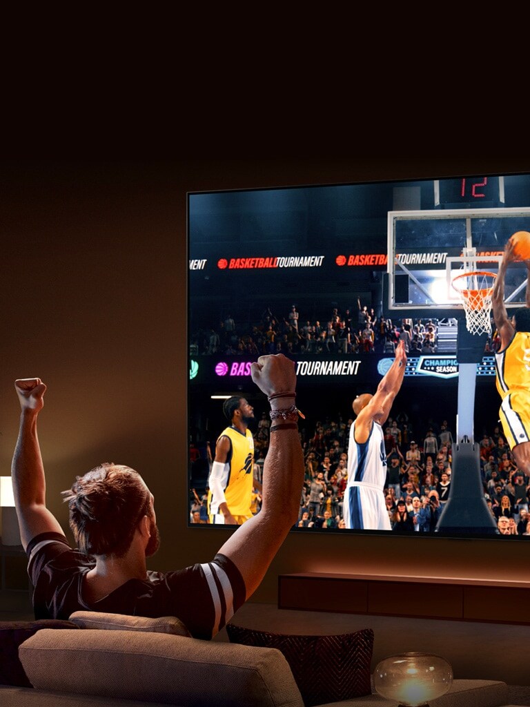 Un homme est assis sur un canapé en face d’une LG TV ultra-large représentant un match de basketball, ses bras étant levés en l’air pour encourager son équipe.