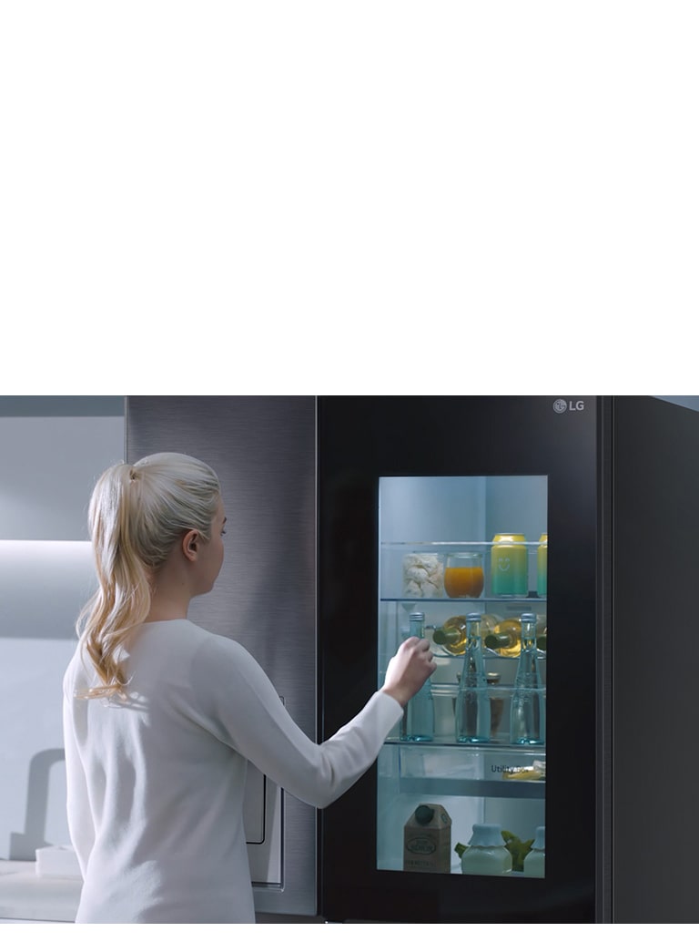 Une vidéo montre une femme qui s’approche de son réfrigérateur InstaView et frappe deux fois. L’intérieur du réfrigérateur s’allume et elle peut voir son contenu sans ouvrir la porte. La vue effectue un zoom avant pour présenter les boissons posées sur la porte, puis un zoom arrière pour voir la femme de derrière alors qu’elle ouvre la porte et prend une boisson.