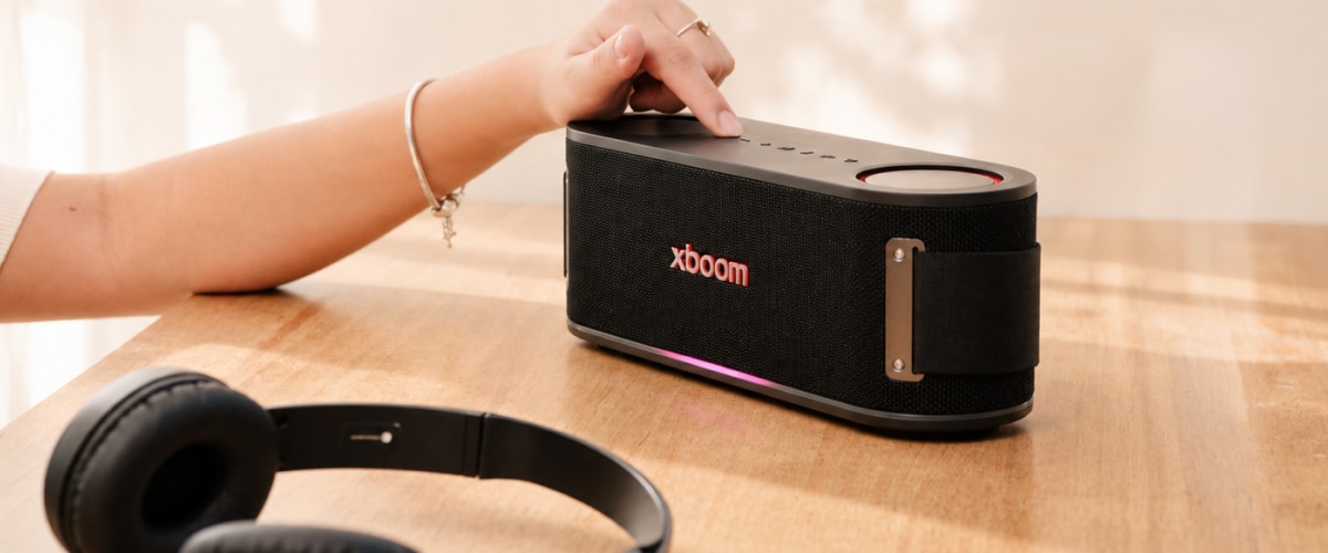 Una mano alcanzando la bocina LG xboom sobre una mesa de madera, con unos audífonos a un lado